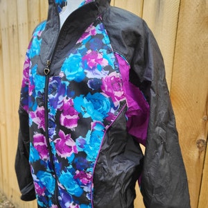 Puede incluir: Una chaqueta cortavientos negra con estampado floral en tonos azules, rosas y morados. La chaqueta tiene cierre de cremallera y cuello alto.