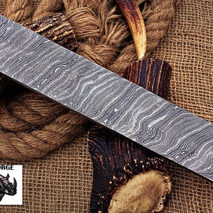 May include: A close-up of a long, rectangular, silver-colored knife blade with a wavy, patterned surface. The blade is lying on a brown, woven fabric background.