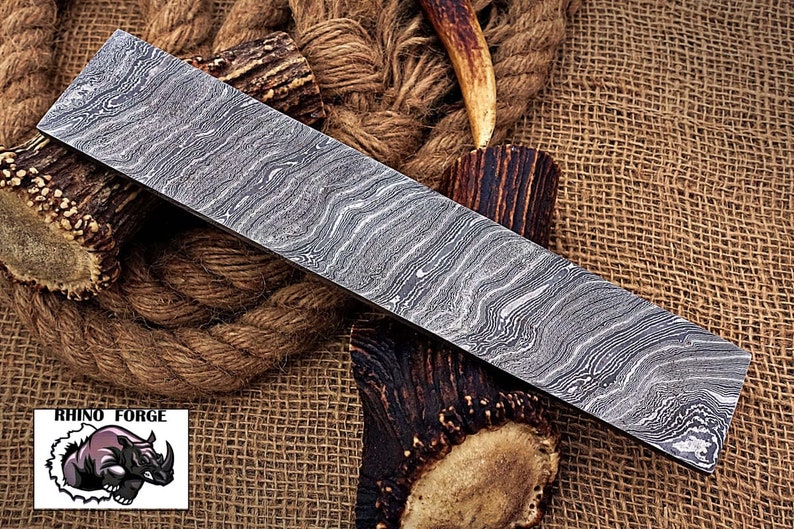 May include: A close-up of a long, rectangular, silver-colored knife blade with a wavy, patterned surface. The blade is lying on a brown, woven fabric background.