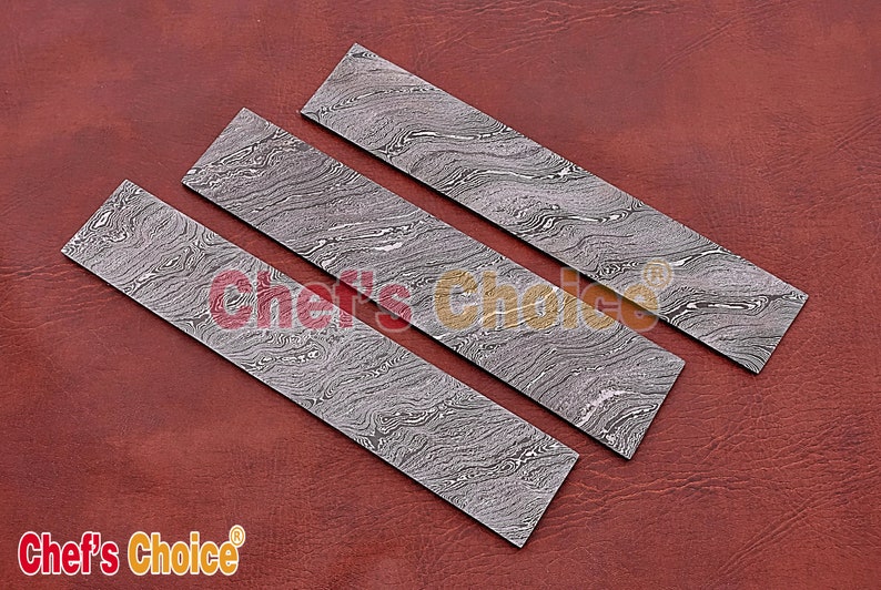 May include: Three rectangular pieces of patterned metal, with a swirling, marbled design in shades of gray. The metal pieces are arranged in a staggered pattern on a brown leather surface.