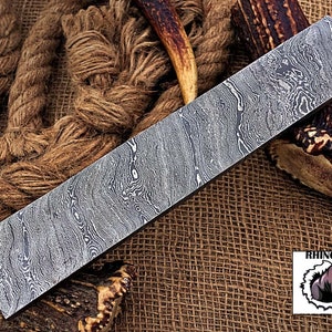 May include: A long, rectangular, silver-colored knife blade with a patterned surface. The blade is lying on a brown burlap surface with a piece of antler on either side. The text "RHINO FORGE" is visible in the lower right corner of the image.