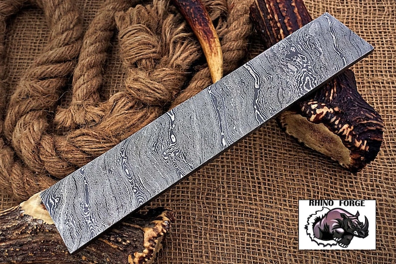 May include: A long, rectangular, silver-colored knife blade with a patterned surface. The blade is lying on a brown burlap surface with a piece of antler on either side. The text "RHINO FORGE" is visible in the lower right corner of the image.