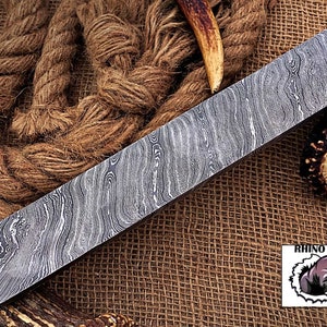 May include: A long, rectangular piece of Damascus steel with a wavy pattern. The steel is lying on a brown burlap surface. The text "RHINO FORGE" is visible in the lower right corner of the image.