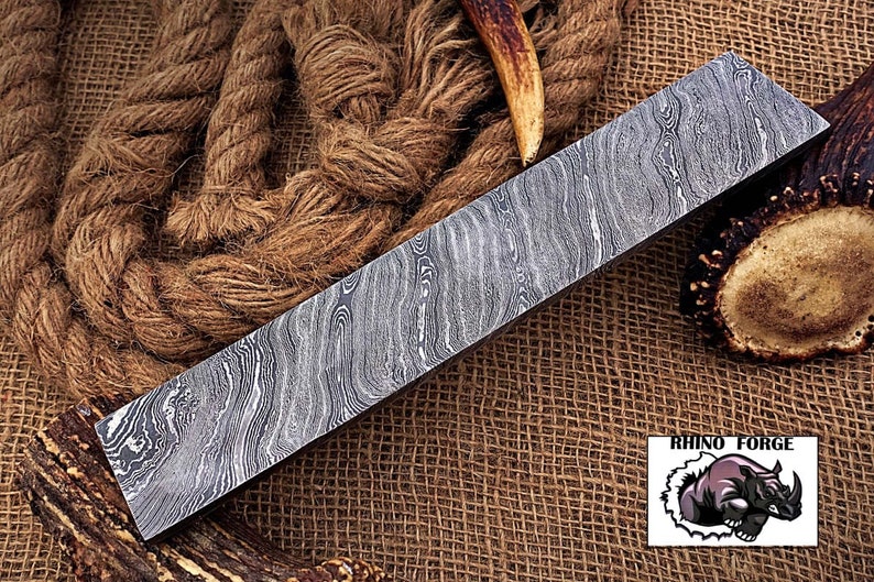 May include: A long, rectangular piece of Damascus steel with a wavy pattern. The steel is lying on a brown burlap surface. The text "RHINO FORGE" is visible in the lower right corner of the image.