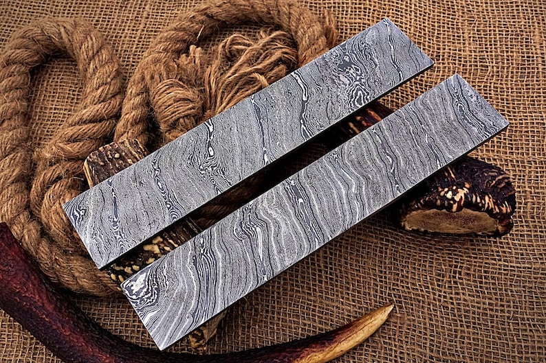 May include: Two long, rectangular pieces of metal with a wavy, patterned surface. The metal is a dark gray color with a silver sheen. The pieces are lying on a brown burlap surface.