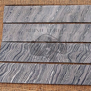 May include: Four pieces of black and white patterned steel knife blanks. The blanks are arranged in a vertical stack on a brown burlap background.