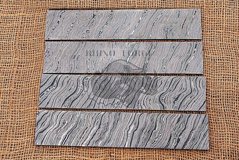 May include: Four pieces of black and white patterned steel knife blanks. The blanks are arranged in a vertical stack on a brown burlap background.