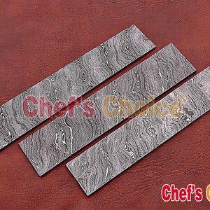 May include: Three Damascus steel knife blanks with a wavy pattern. The blanks are arranged in a row on a brown leather surface.  Chef's Choice&reg; is printed on the surface.