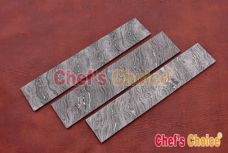 May include: Three Damascus steel knife blanks with a wavy pattern. The blanks are arranged in a row on a brown leather surface.  Chef's Choice&reg; is printed on the surface.