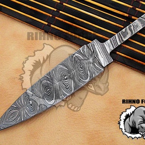 May include: A Damascus steel knife blade with a unique wood grain pattern. The blade has a pointed tip and a wide, flat body. The image includes the text "RIHNO FORGE" and a rhino logo.