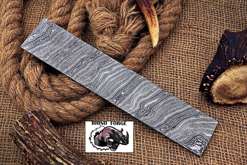 May include: A long, rectangular piece of Damascus steel with a wavy pattern. The steel is a light gray color with darker gray lines running through it. The steel is sitting on a brown burlap background. The text "RHINO FORGE" is visible in the lower center of the image.