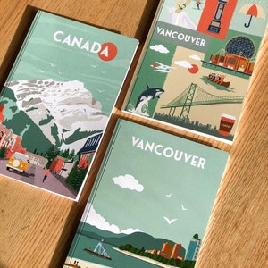 May include: Three notebooks with travel-themed illustrations. One says "CANADA" with a mountain scene. Two say "VANCOUVER" with city and nature illustrations, including a bridge, orca, and clock tower. The notebooks are on a wooden surface.