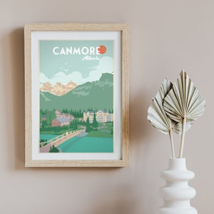 May include: A vintage-style travel poster with the text "Canmore, Alberta" in a blue and white font. The poster features a scenic view of a town with mountains in the background and a bridge over a lake.