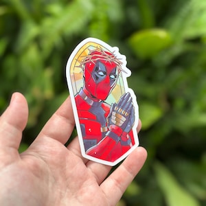 May include: A sticker featuring a cartoon depiction of Deadpool wearing a crown of thorns and praying with his hands clasped together. The background is a stained glass window with a halo around Deadpool's head.