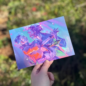 Op de afbeelding: Een print met een levendige illustratie van twee dansende figuren, weergegeven in tinten paars, oranje en blauw. Het kunstwerk heeft een dynamische compositie met de tekst "JENNIE PLANET" in de hoek.