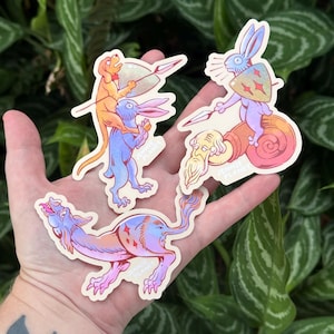 May include: A hand displays three quirky stickers. One shows a dog atop a rabbit, another a rabbit riding a snail, and the third a mythical beast. Each sticker has a unique design with bright colours and detailed artwork.