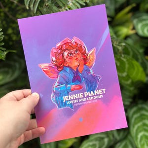 May include: A print with an illustration of a person with red hair, glasses, and wings, wearing a blue jacket and a pink hat. The print has the text "JENNIE PLANET ARTIST AND TATTOOIST @JENNIEPLANETINK" on a purple and orange background.
