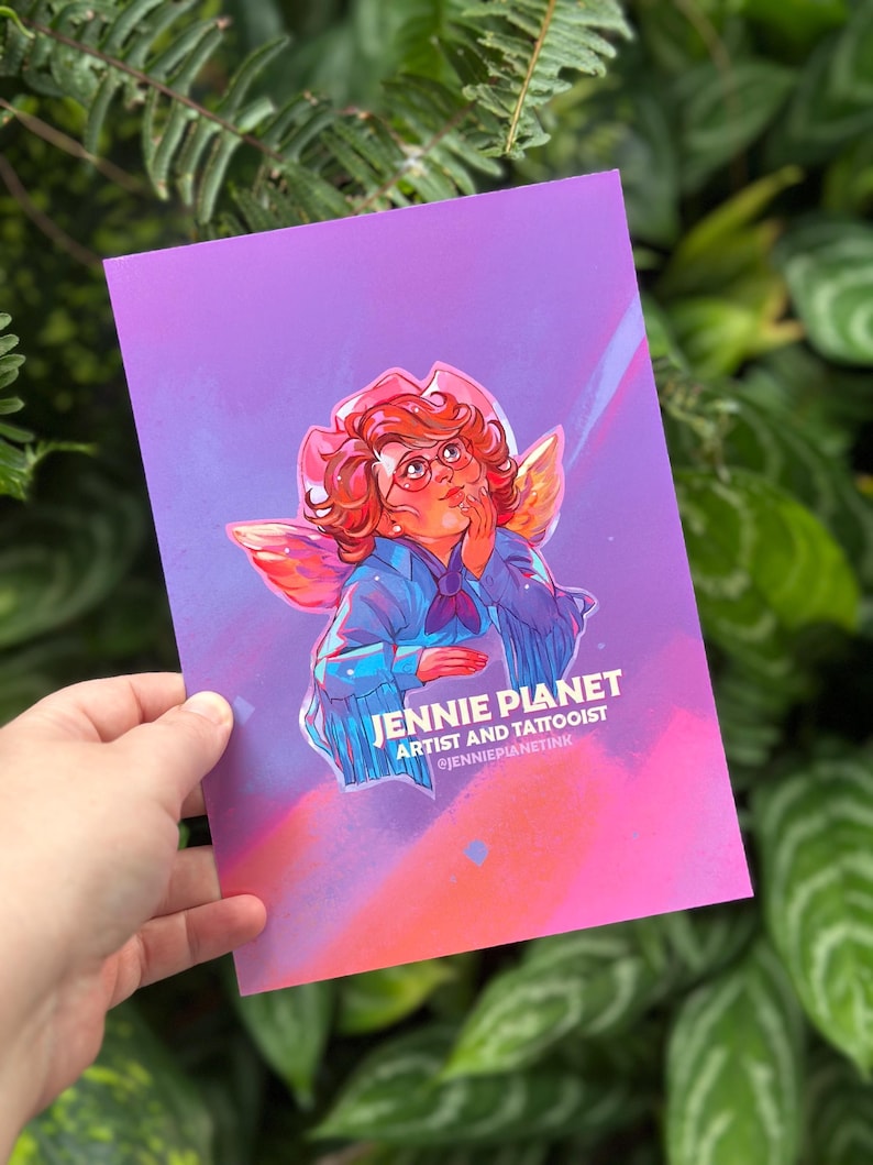 May include: A print with an illustration of a person with red hair, glasses, and wings, wearing a blue jacket and a pink hat. The print has the text "JENNIE PLANET ARTIST AND TATTOOIST @JENNIEPLANETINK" on a purple and orange background.