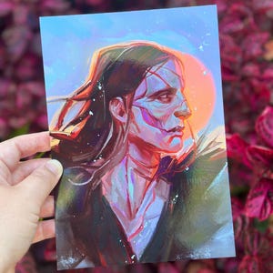 May include: A portrait print depicting a person with long dark hair and a side profile. The artwork uses blues, purples, and oranges, with a bright orange sun-like shape in the background. The person's face has visible brushstrokes.