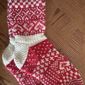 May include: A pair of knitted socks with a red and white geometric pattern. The socks have a white cuff and heel, and the red sections feature a repeating white design. The socks are made of a soft, warm material.