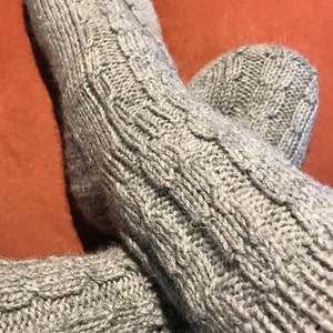 May include: A pair of gray knitted socks with a textured cable knit pattern. The socks are made of a soft, warm material, perfect for cold weather. The socks are shown on a red-orange surface.