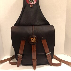 May include: A brown leather saddlebag with a lion head design on the flap. The bag has a zippered pocket on the front and a buckle closure. The bag is made of high-quality leather and has a rustic look.