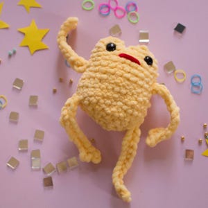 Peut inclure: Une grenouille au crochet jaune clair avec des yeux noirs et une bouche rouge. La grenouille est entourée d'étoiles jaunes, de petits miroirs carrés, d'élastiques colorés et de perles dorées sur un fond violet clair. Un jouet fantaisiste fait à la main.