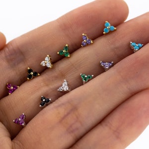 以下が含まれることがあります： 三角形の小さなイヤリングが10個、それぞれに小さな宝石が3つずつ付いています。イヤリングは手に置かれており、それぞれ異なる色の宝石が付いています。ターコイズ、パープル、グリーン、ブラック、ホワイト、ピンク、レッドです。