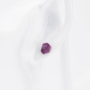 May include: A close-up of a purple gemstone earring stud. The earring is set in a silver setting and is worn in a person's ear.