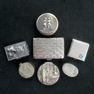 May include: A collection of seven silver-toned metal boxes and cases of various shapes and sizes. The items feature diverse embossed designs including a pair of figures, a brick pattern, grape clusters, a building, and floral accents on a textured surface.