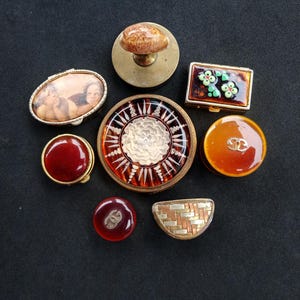 May include: A collection of eight decorative items including a brass stamp, an oval box with a cherub image, a rectangular box with floral accents, a large circular medallion with a white flower, and several round and semi-circular buttons.