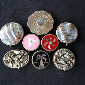 Pode incluir: Uma coleção de broches vintage com vários designs. Os broches apresentam materiais como concha de abalone, pirite e esmalte nas cores prata, rosa, vermelho e preto. Os broches são redondos e ovais, com fechos de alfinete.