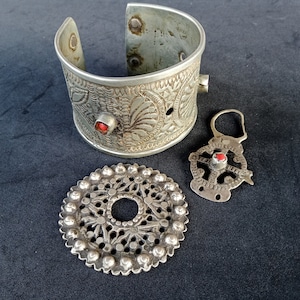 May include: Three silver jewelry pieces with red gemstone accents. A wide silver bracelet with an intricate floral design, a round silver pendant with a cutout design, and a silver charm with a hook.
