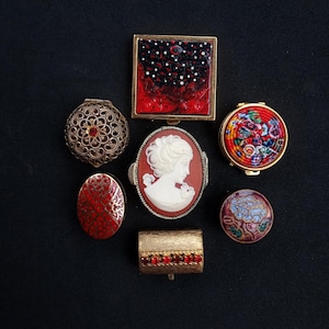 Op de afbeelding: Een verzameling van zeven kleine decoratieve doosjes en poederdoosjes. De items omvatten een vierkant zwart en rood doosje, een rond filigraan doosje met een rode steen, een rond bloemrijk poederdoosje, een ovaal rood patroondoosje en een camee.