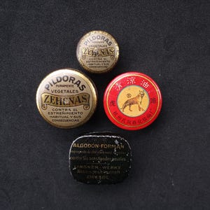 May include: Four round metal tins with different labels. The first tin is gold with black text that reads "PILDORAS PURAMENTE VEGETALES ZEHNAS CONTRA EL ESTREÑIMIENTO HABITUAL Y SUS CONSECUENCIAS". The second tin is gold with black text that reads "PILDORAS PURAMENTE VEGETALES ZEHNAS CONTRA EL ESTREÑIMIENTO HABITUAL Y SUS CONSECUENCIAS". The third tin is red with gold text that reads "虎跑优州孔容化妆品". The fourth tin is black with white text that reads "ALGODON-FORMAN empeados de aire claro para limpiar contra los constipados pasales. LINGNER WERKE Aktiengesellschaft DRESDE".