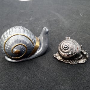 Puede incluir: Dos figuras de caracol. La más grande es plateada con una banda dorada alrededor de la concha. La más pequeña es una caja con bisagras, también con forma de caracol, con un diseño detallado de la concha.