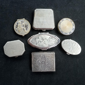 Puede incluir: Una colección de siete cajas pequeñas de metal en varias formas, incluyendo redondas, ovaladas, rectangulares y octogonales. Las cajas presentan diversas texturas superficiales, incluyendo patrones grabados intrincados, diseños geométricos y escenas figurativas en relieve.