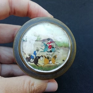 Könnte beinhalten: Runde, antike Schmuckdose mit detaillierter Miniaturmalerei von Reitern auf Pferden. Das Gemälde zeigt Figuren in farbenfroher Kleidung auf Pferden verschiedener Farben, vor einer Landschaft. Die Dose hat einen dunklen, gealterten Metallrahmen.