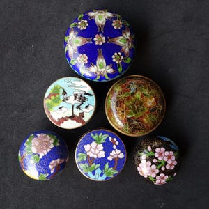 Op de afbeelding: Een verzameling van zes kleine, ronde, decoratieve doosjes met ingewikkelde emaille ontwerpen. De doosjes hebben bloemen- en vogelmotieven in verschillende kleuren, waaronder blauw, groen, roze en goud. De deksels zijn gewelfd en hebben een metallic gouden rand.