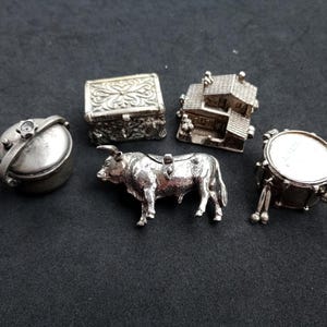 Puede incluir: Una colección de cinco figuras de metal en miniatura de tono plateado que incluye una olla redonda con tapa, una caja rectangular ornamentada, una casa pequeña, un toro y un tambor con dos baquetas. Cada pieza presenta texturas detalladas.