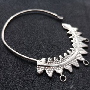 May include: A silver earring component with a half-circle design. The earring features a decorative, textured section with triangular shapes and small loops for attachment. The earring is shown against a dark background.