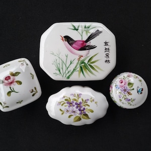 May include: Four white porcelain trinket boxes with hand-painted designs. The largest box has a pink and black bird on a bamboo branch. Other boxes feature floral patterns in pink, purple, and blue. The boxes are decorative.