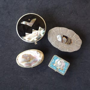 Puede incluir: Cuatro broches decorativos con incrustaciones de concha de abulón iridiscente. Los broches tienen varias formas: un círculo con fondo negro, un óvalo, una caja rectangular con detalles turquesa y un óvalo con borde plateado.