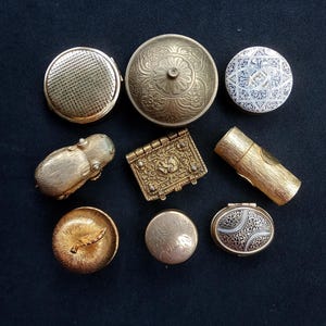 May include: A collection of nine small metal containers and trinket boxes in various shapes, including round, oval, and rectangular forms. The items feature gold-toned finishes, intricate embossed floral patterns, geometric engravings, and textured surfaces.