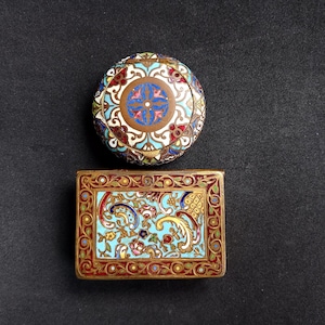 Puede incluir: Dos cajas antiguas y ornamentadas con intrincados diseños de esmalte. La caja redonda presenta un motivo central de cruz azul, mientras que la caja rectangular muestra patrones florales en azul, rojo y dorado. Ambas sobre un fondo oscuro.