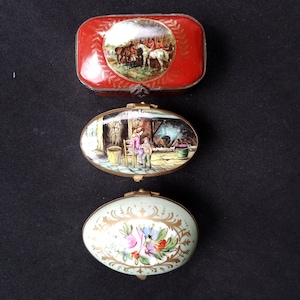 May include: Three antique decorative trinket boxes. The top box is rectangular and red with a painting of people and horses. The middle box is oval with a scene of people near a fire. The bottom box is oval and light green with floral designs.