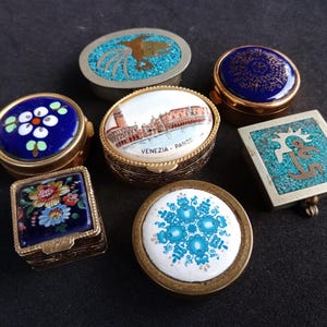 May include: Seven small decorative boxes with various designs. The boxes are made of metal and have a gold finish. The lids are decorated with colorful enamel and feature images of flowers, a bird, a city scene, and a geometric pattern. The boxes are all closed.