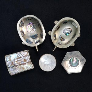 Puede incluir: Un surtido de piezas de joyería plateadas, que incluyen dos cuencos con forma de tortuga, un cuadrado, un redondo y una pieza hexagonal. Cada artículo presenta incrustaciones de concha de abulón iridiscente, creando un efecto brillante sobre fondo oscuro.