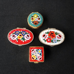 May include: Four mosaic trinket boxes in various shapes: oval, round, and square. Each box features a floral design with red, white, blue, and yellow flowers. The boxes have gold-coloured metal frames and are set against a black background.