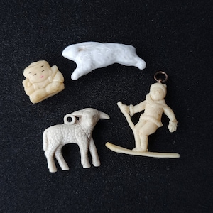 Puede incluir: Una colección de pequeñas figuras blanquecinas sobre una superficie negra. Las figuras incluyen un cordero, un esquiador, una figura sentada y un conejo. Cada figura tiene un lazo para colgar. El esquiador sostiene bastones de esquí.